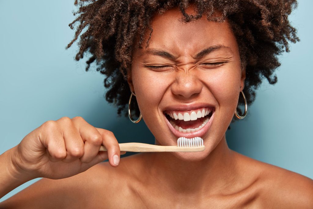 Lächelnde Frau mit geöffneten Mund und geschlossenen Augen, die eine Bambuszahnbürste hält, Dr. Bruhn Zahnarztpraxis Berlin-Süd